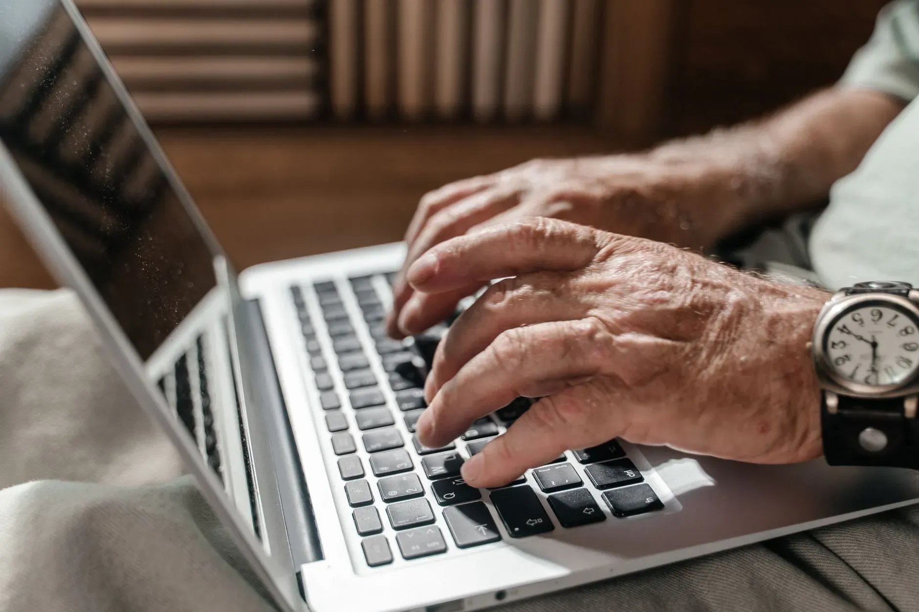 AI25.Studio AI GENERATIVE Primo piano delle mani di una persona anziana che digita su un computer portatile e indossa un orologio da polso, in un ufficio a casa.